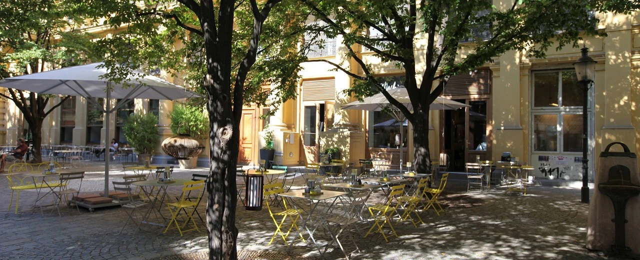 © Mochi Gemütliche Café-Terrasse mit Sonnenschirmen, Bäumen & gelben Stühlen. Perfekt zum Entspannen.