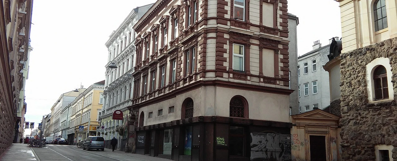 © Marion Ramell Enge Gasse mit Jugendstil-Gebäuden. Beige, weiße & braune Fassaden, Autos & Fahrräder.