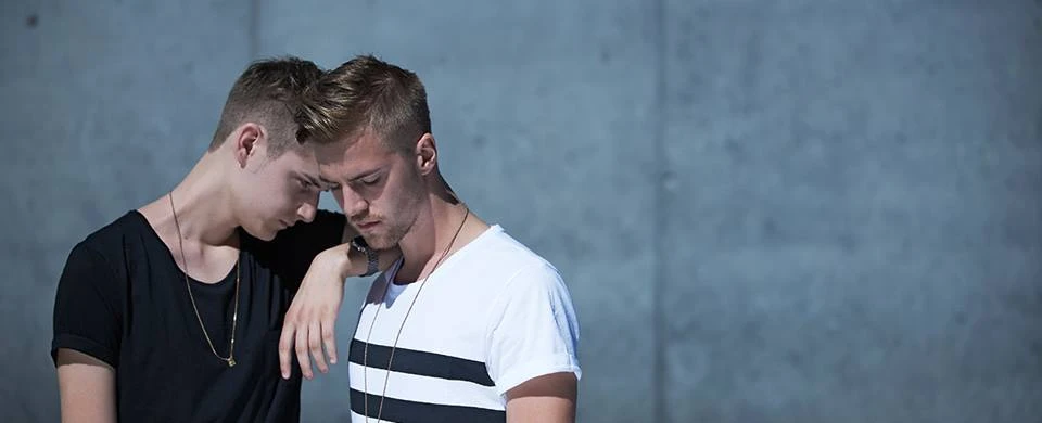 Zwei junge Männer, T-Shirts tragend, stehen nah beieinander vor Betonwand. Nachdenkliche Stimmung.