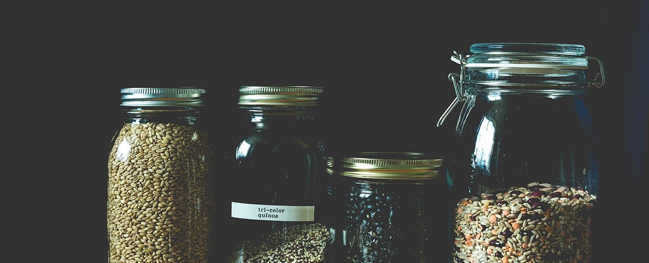 Vier Gläser mit verschiedenen Körnern und Hülsenfrüchten (Quinoa, Sonnenblumenkerne, schwarze Bohnen, Getreidemischung) auf dunklem Hintergrund.