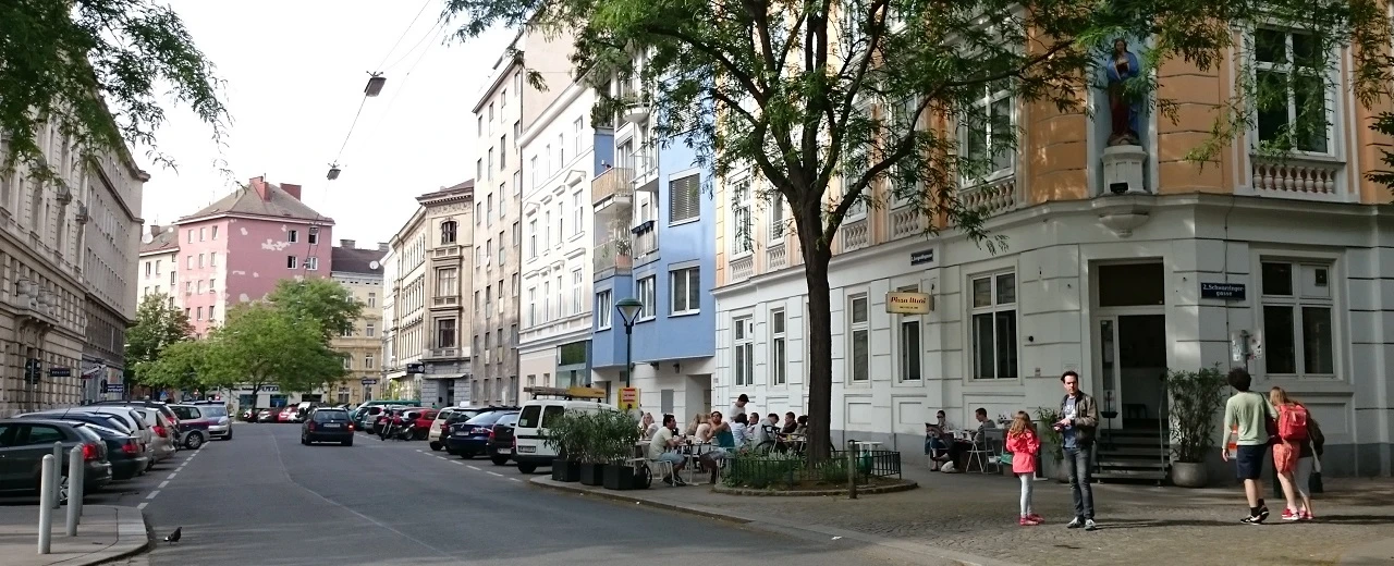 © Miriam Kummer Stadtstraße mit Gebäuden, geparkten Autos & Menschen vor Restaurant. Friedliche Alltagsszene.