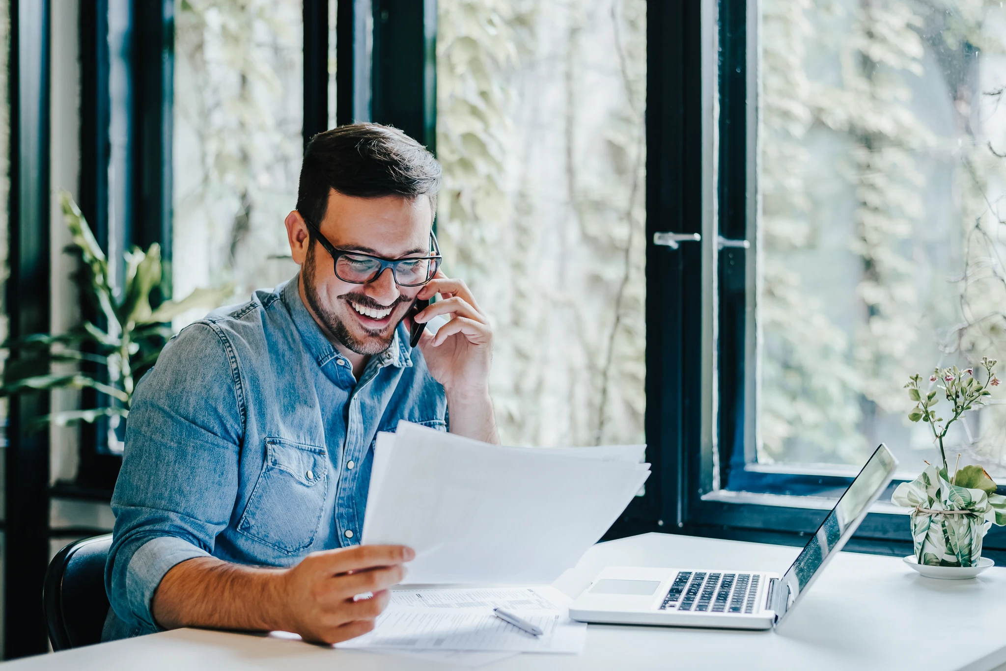 Mann sitzt lachend mit Unterlagen über Immobilien Investments und Laptop am Schreibtisch und telefoniert