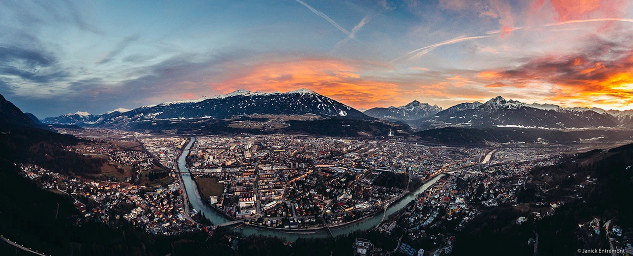 Panoramabild von Innsbruck bei Sonnenuntergang. Die Stadt liegt in den Alpen, ein Fluss schlängelt sich durchs Zentrum. Berge sind schneebedeckt und in warmen Farben des Sonnenuntergangs getaucht.