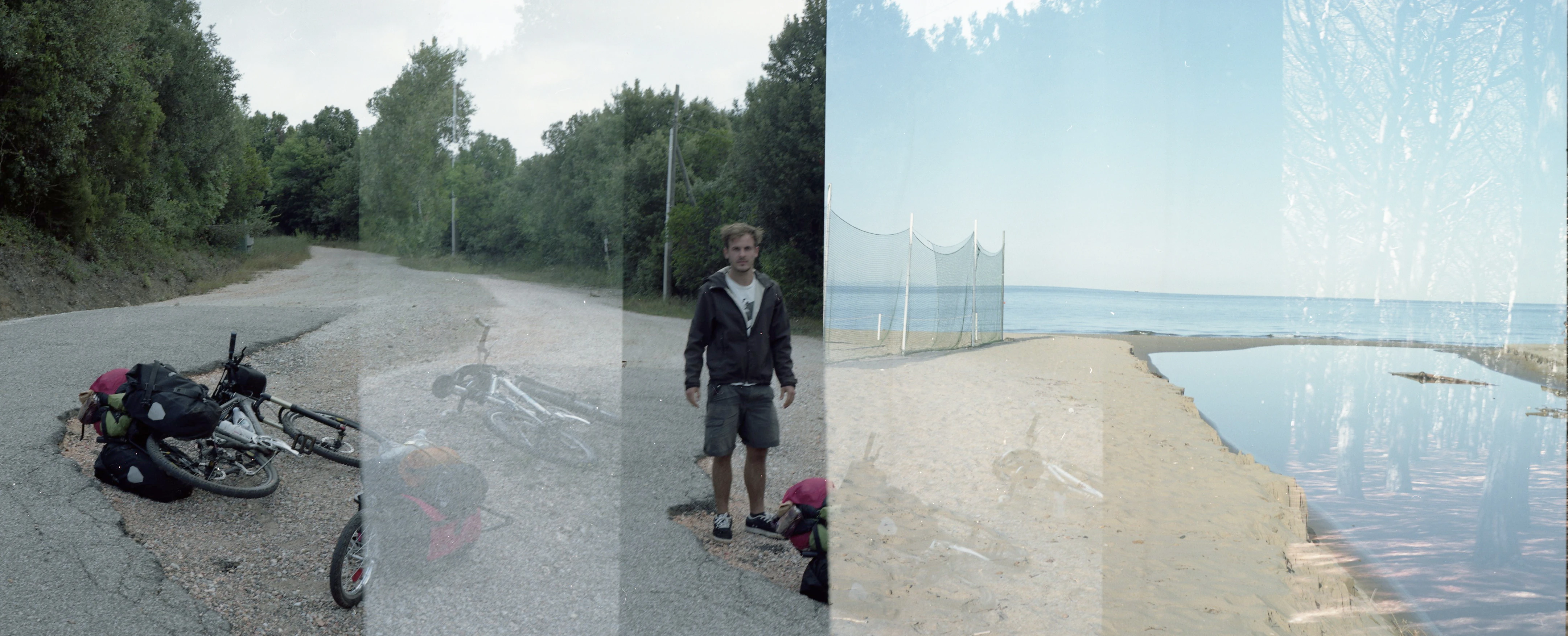 © Aslan Kudrnofsky Radfahrer mit Gepäck am Strand, Fahrrad auf kaputter Straße, Meer im Hintergrund. Überlagerungseffekt.