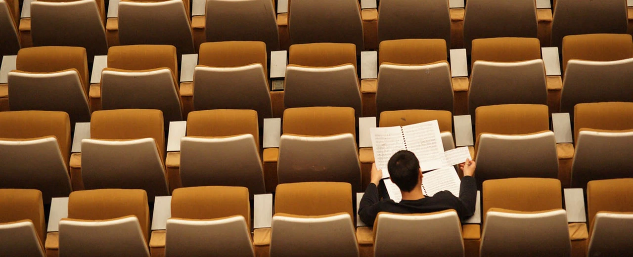 Person liest Noten in leerem Auditorium; beige/goldene Stühle.