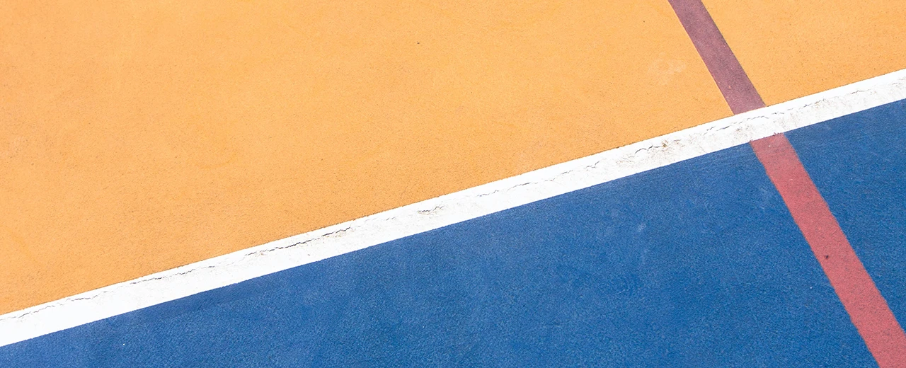 Nahaufnahme eines Sportplatzbodens mit orangenen und blauen Feldern, getrennt durch eine weiße Linie.  Rote Linien sind ebenfalls zu sehen.