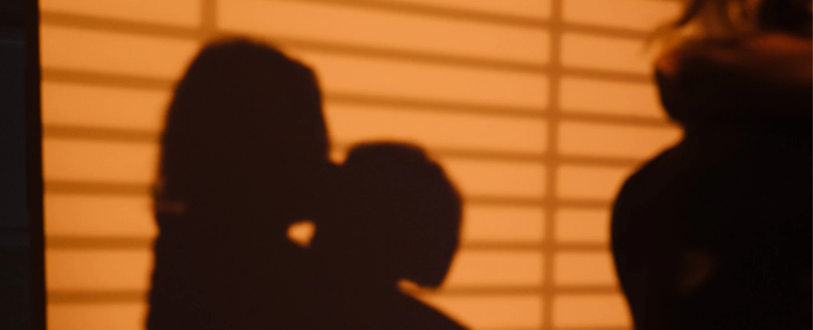 Unscharfe Schatten zweier Personen, die sich vor orangefarbenen, horizontalen Streifen umarmen oder küssen.