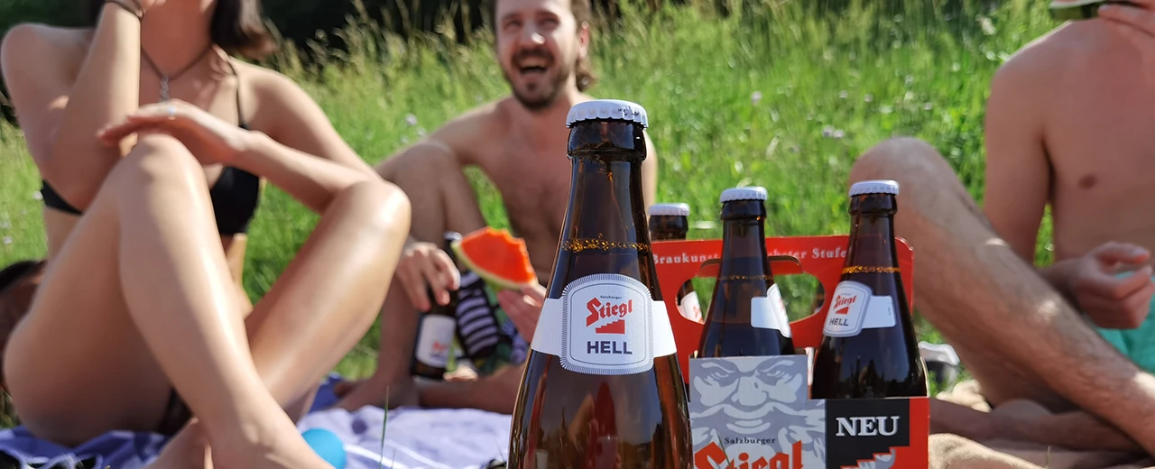 Stiegl Hell Bierflaschen im Vordergrund eines sommerlichen Picknicks mit Freunden.  Erstellt mit ELS-N29.