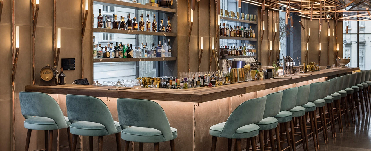 Moderne Bar mit Holztheke, türkisfarbenen Hockern & Kupferakzenten. Viele Spirituosen.