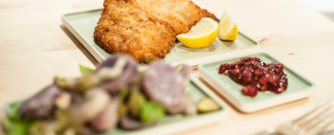 Appetitliches Foto: Paniertes Schnitzel mit Kartoffelsalat, grünen Bohnen und Preiselbeersauce auf quadratischen Tellern.