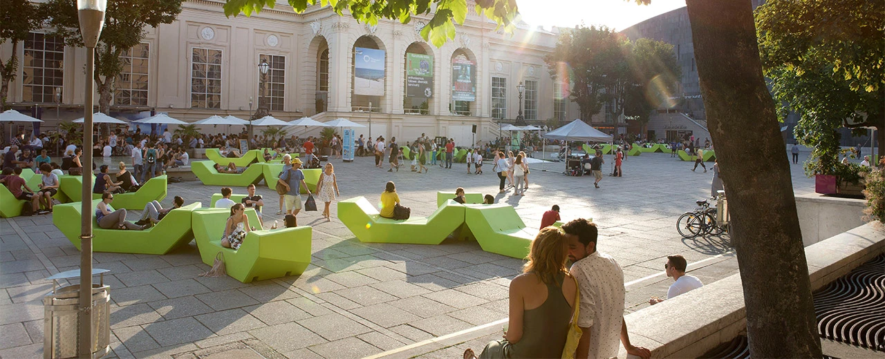 Lebhafter Platz mit limefarbenen Sitzmöbeln, Menschen und einem Gebäude im Hintergrund. Bearbeitet mit Adobe Photoshop Camera Raw.