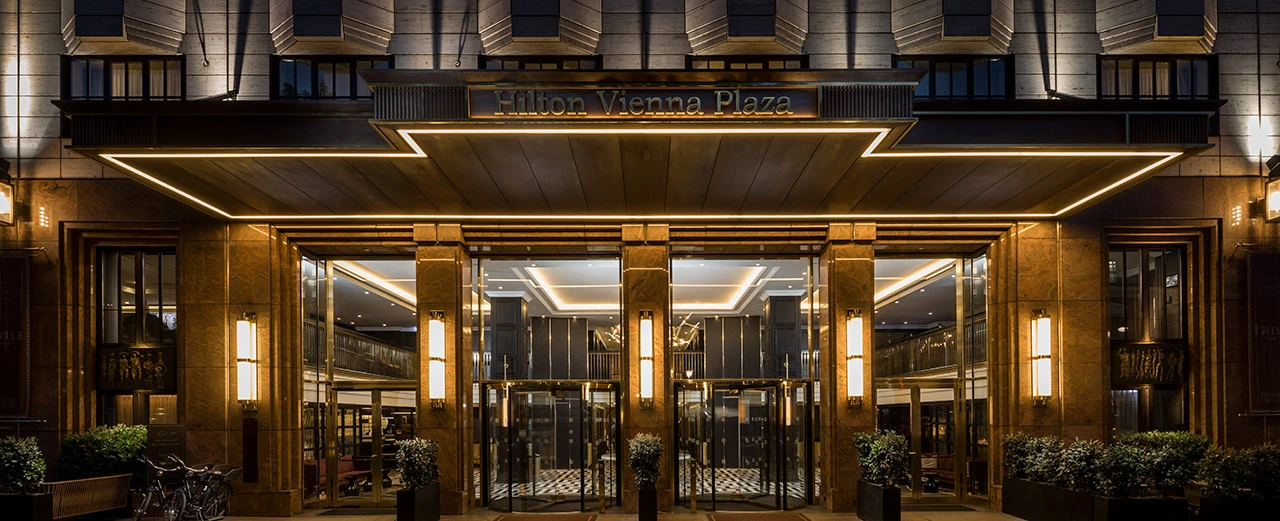 Nachtansicht des eleganten Haupteingangs des Hilton Vienna Plaza in Wien. Art-Deco-Architektur mit Marmor und goldenen Akzenten.