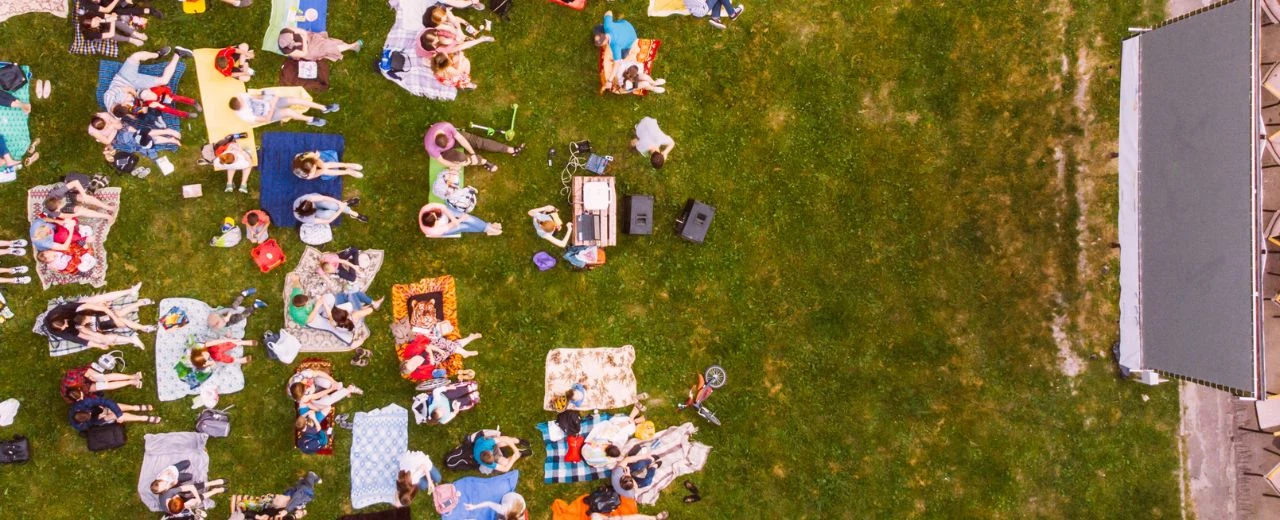 Draufsicht auf ein Open-Air-Kino mit vielen Zuschauern auf Decken im Grünen vor einer großen Leinwand.