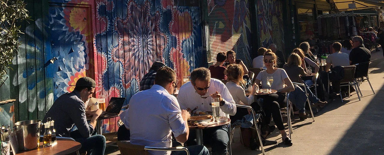 Sonnige Außenterrasse mit Menschen, die an Tischen sitzen und essen und trinken, vor einer bunt bemalten Wand.