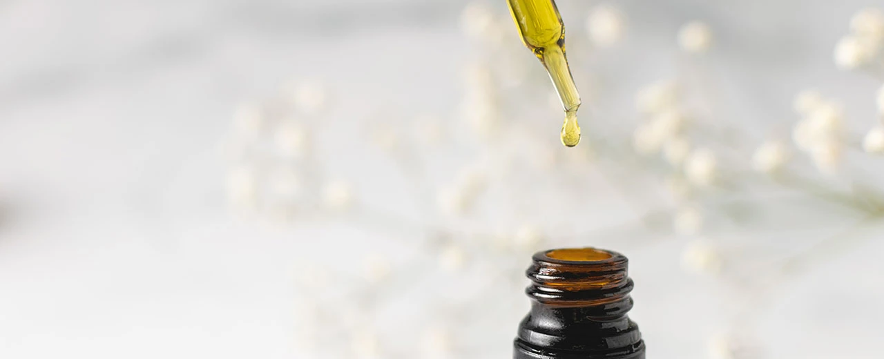 Tropfen ätherisches Öl in Glasflasche, weiße Blüten im Hintergrund. Naturkosmetik, Hautpflege.