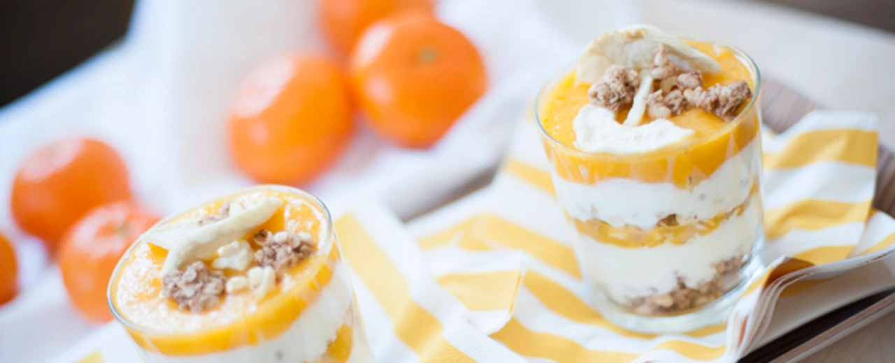 Zwei schmackhafte Dessertschichten in Gläsern mit Joghurt, Orangen und Granola. Mandarinen liegen daneben.