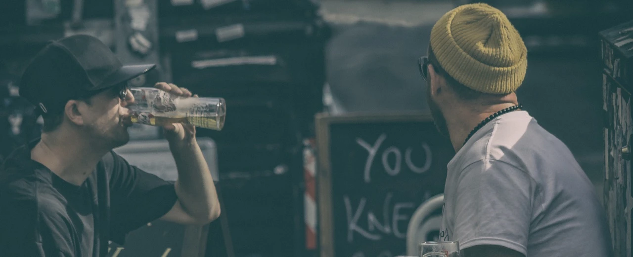 Zwei Männer unterhalten sich draußen, einer trinkt Bier.