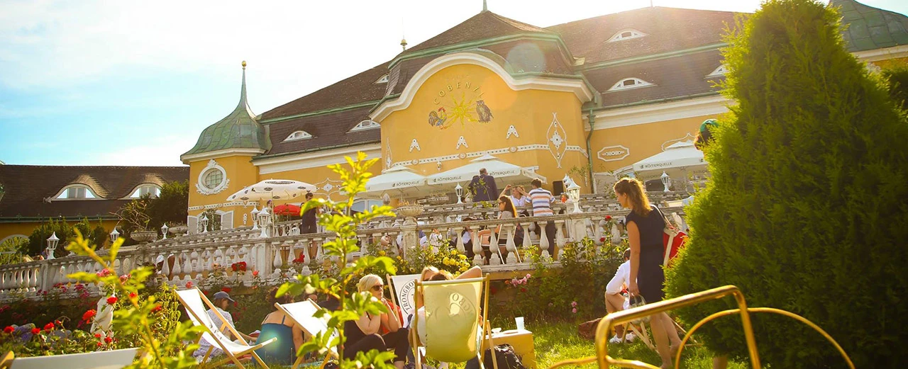 Idyllisches Bild von einem gelben Gebäude mit grünem Dach und Terrasse, umgeben von Rosen und entspannten Gästen. Schöner Garten und sonnige Atmosphäre.
