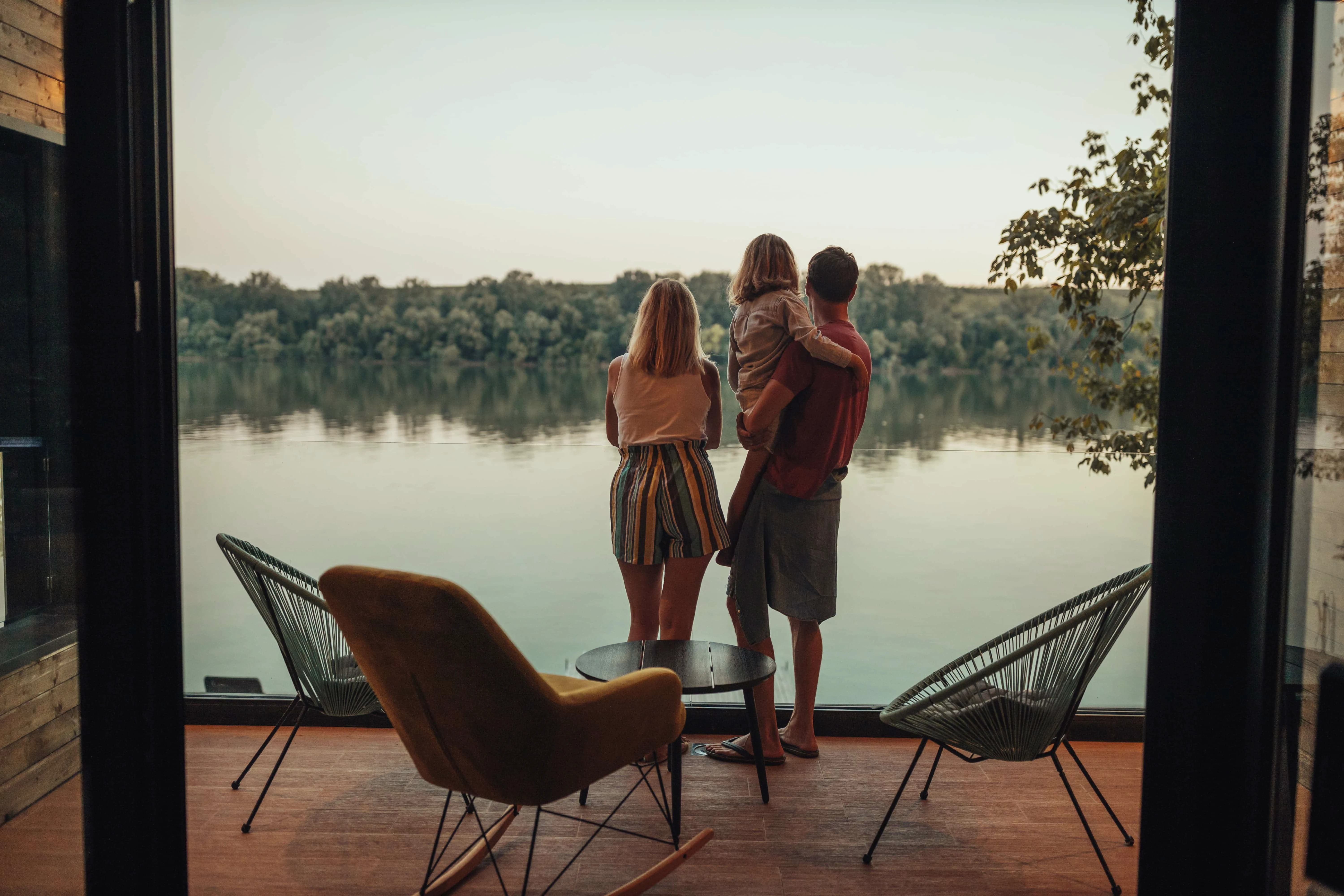 Familie mit Kind blickt in der Dämmerung von Ihrer Holzterrasse auf den See
