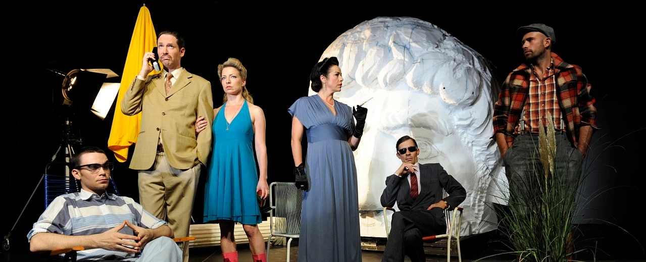 Schauspieler in Retro-Kostümen bei einer Theateraufführung vor einer großen Skulptur.