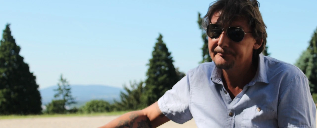 Mann mit Sonnenbrille vor Bäumen, hellblaues Hemd, Tattoo.