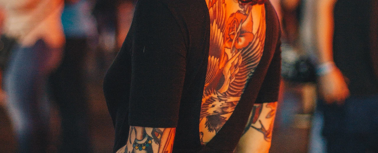 Tattoo auf dem Rücken einer Person mit Flügeln und einer Rose. Schwarzes Shirt, unscharfer Hintergrund.