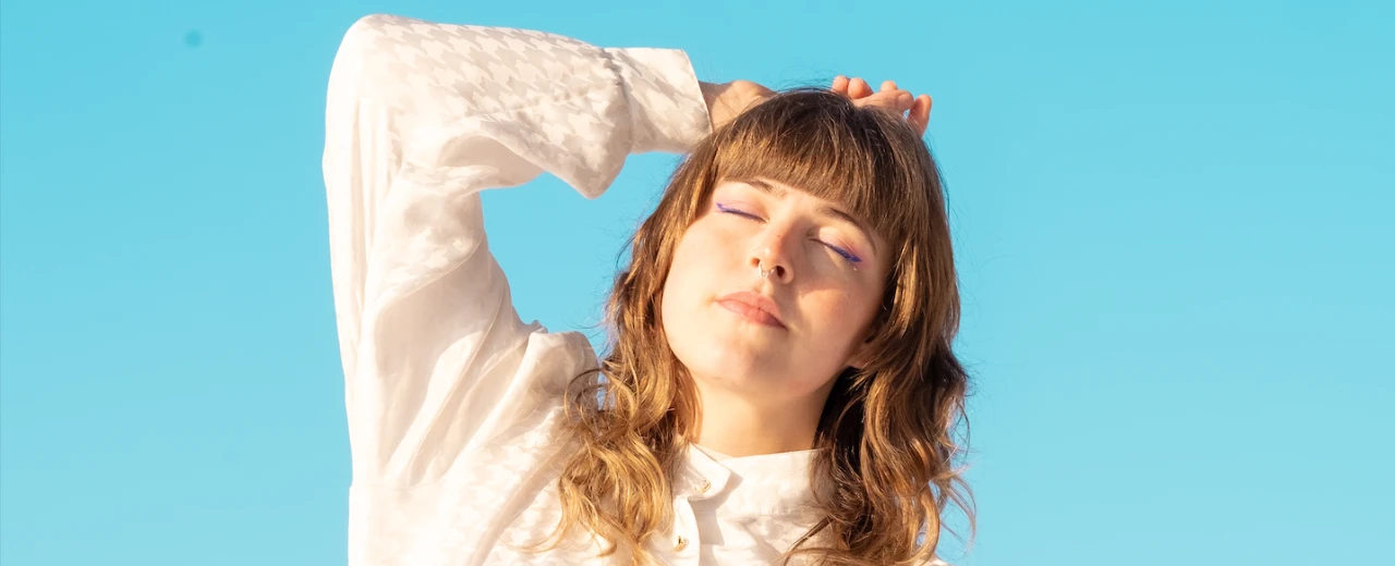 Junge Frau mit geschlossenen Augen, braunen Haaren und weißem Oberteil vor hellblauem Himmel.