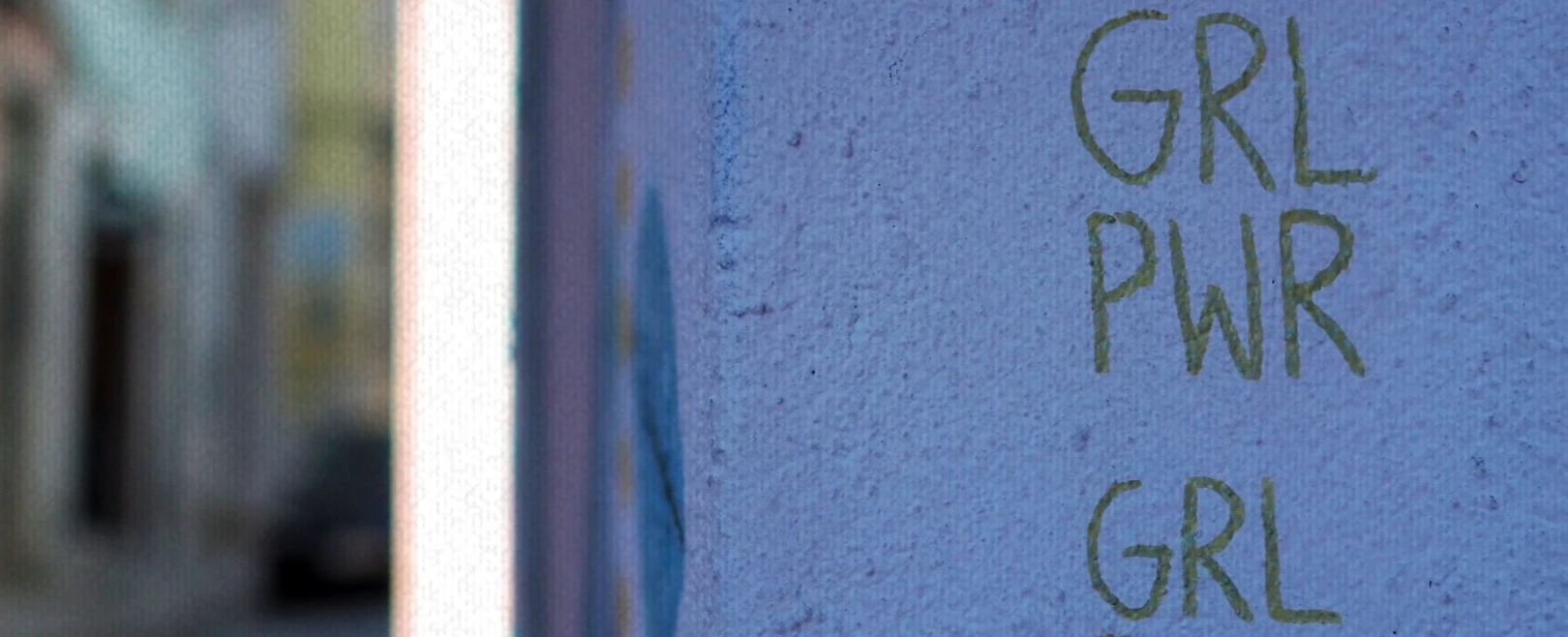 Graffiti an einer Wand: 'GRL PWR GRL' in Gelb auf lilafarbenem Hintergrund.