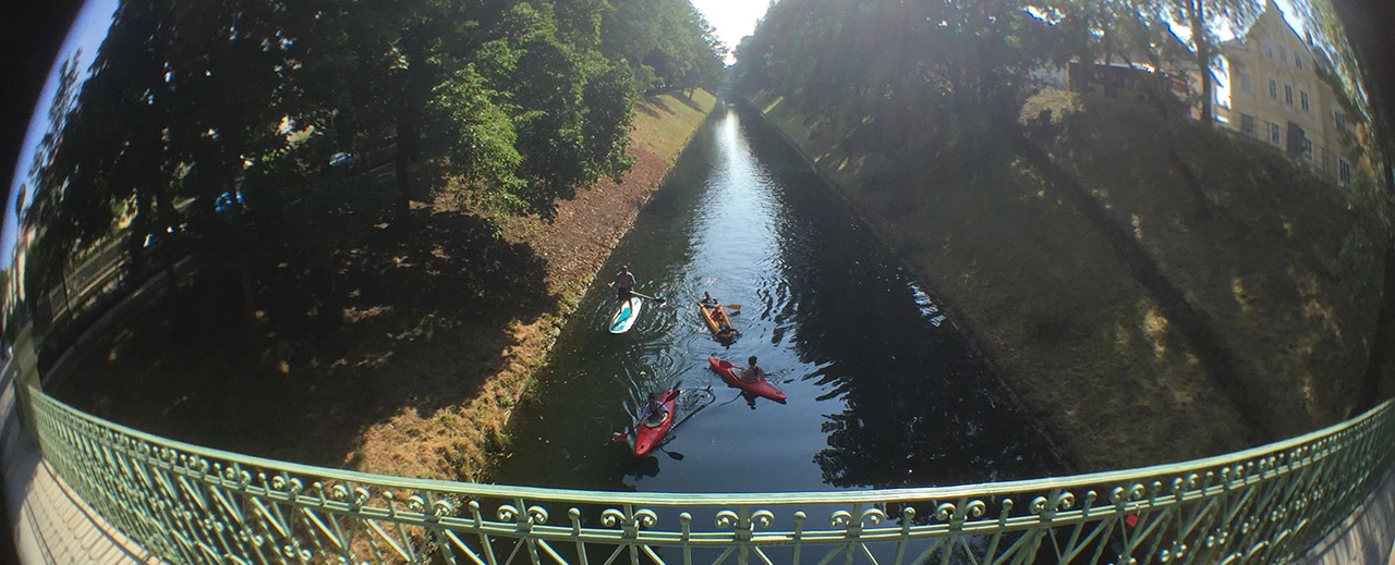Kajaks & SUP-Boards auf ruhigem Kanal, von Brücke mit grünem Geländer gesehen.