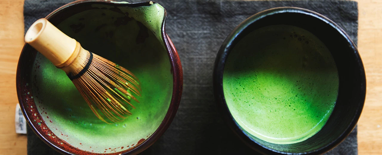 Matcha-Tee wird in einer Schale mit einem Bambusbesen aufgeschlagen. Daneben steht eine weitere Schale mit fertigem Matcha.