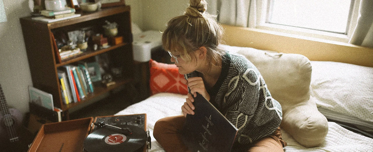 © Unsplash Frau hört entspannt Schallplatte auf dem Bett. Gemütliche Atmosphäre mit Büchern und Vintage-Details.