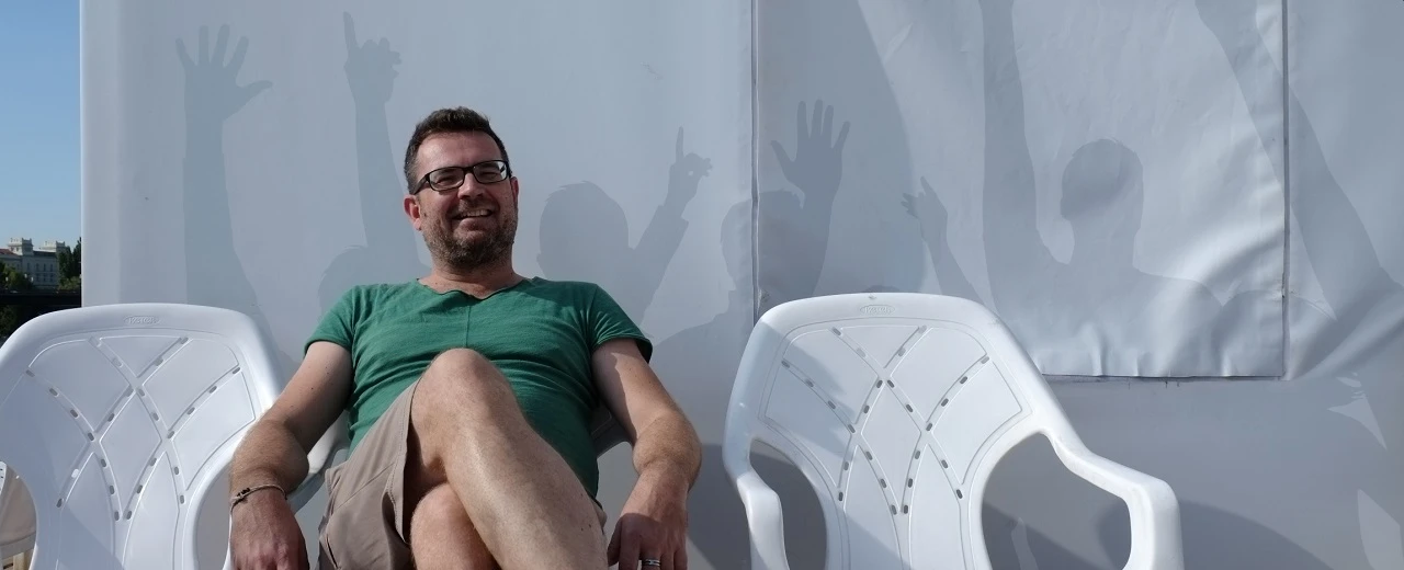 Lächelnder Mann sitzt auf weißen Plastikstühlen vor einer Wand mit Schatten von Händen.