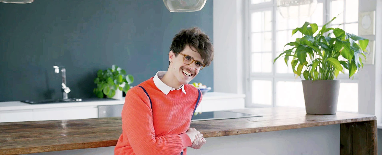 Lächelnder Mann in orangem Pullover in moderner Küche mit Holzarbeitsplatte.