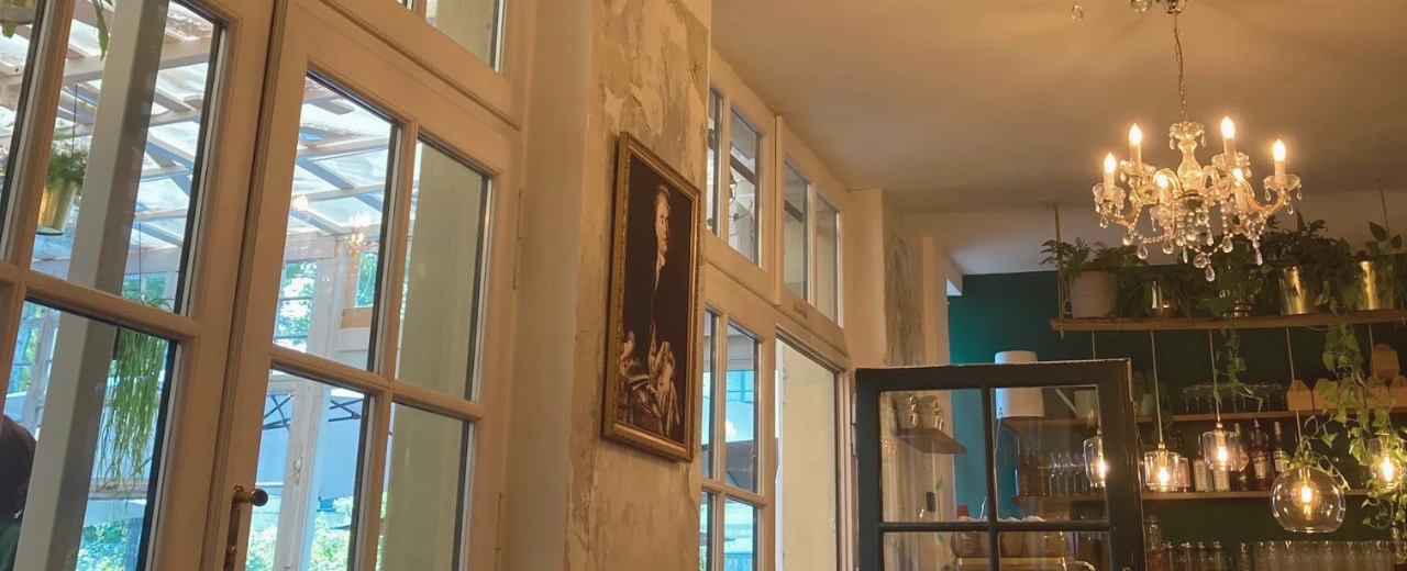 Stilvolles Café-Interieur mit großen Fenstern, antikem Gemälde, Kronleuchter und Pflanzen. Gemütliche und elegante Atmosphäre.