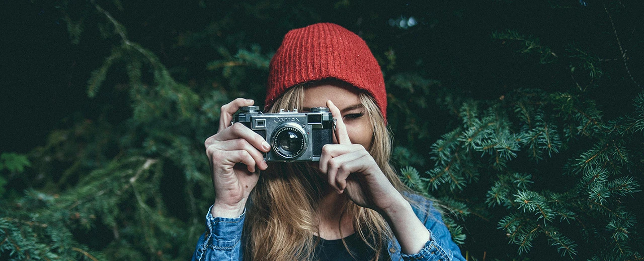Junge Frau mit roter Mütze fotografiert mit einer Vintage-Kamera in einem dunklen Wald.