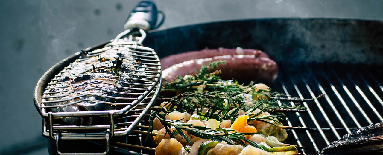 Leckeres Grillgut: Fisch im Korb, Würstchen und Hähnchenspieße mit Gemüse auf einem heißen Grill.