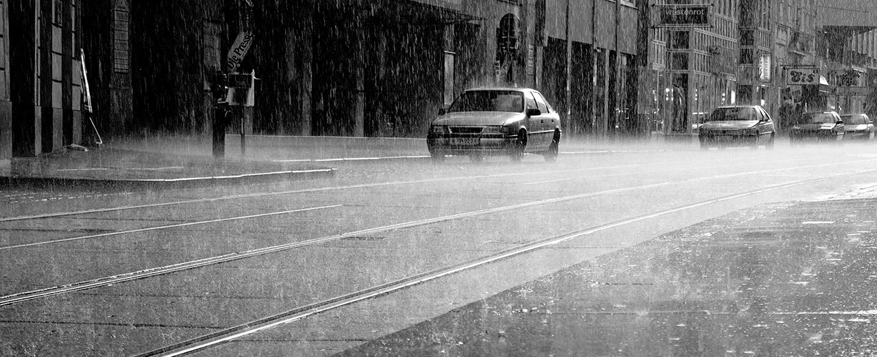 Schwarz-weiß Foto: Stadtstraße bei starkem Regen, Straßenbahnlinie, mehrere Autos.