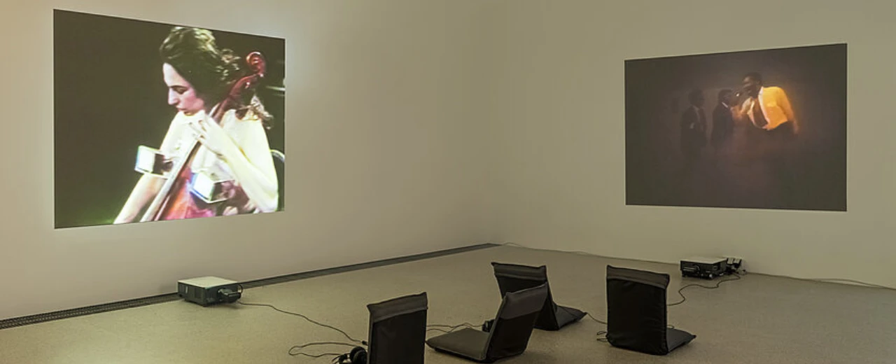© Mumok/Klaus Pichler Moderne Kunstinstallation: Zwei Projektionen (Cellistin, Gruppenszene), drei Stühle, Projektoren. Videoinstallation, Multimedia-Kunst.