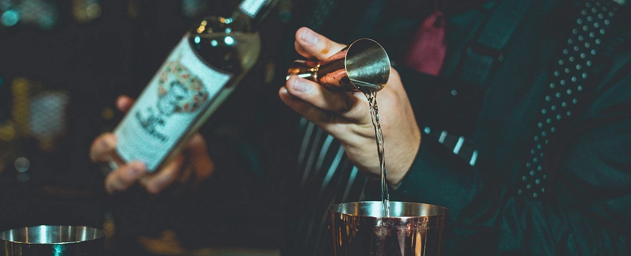© Ivan Cortez via Unsplash Barkeeper gießt Spirituose in Cocktailshaker. Nahaufnahme Hände & Utensilien.