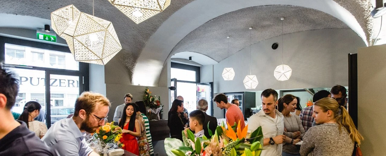 Eröffnungsfeier: Modernes Lokal mit vielen Gästen, geometrischen Lampen & Blumen. Freundliche Atmosphäre.