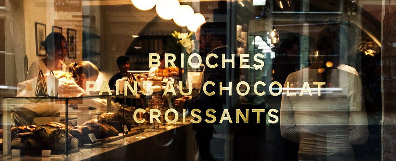 © Paremi, Charlotte Forest Schaufenster einer Bäckerei mit goldenen Schriftzügen: Brioches, Pains au Chocolat, Croissants.  Mitarbeiter und Backwaren im Hintergrund.