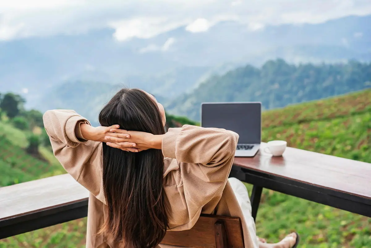 Eine junge Frau genießt ihre Workation: Sie arbeitet an ihrem Laptop, während sie die Ausblick auf die Berge genießt