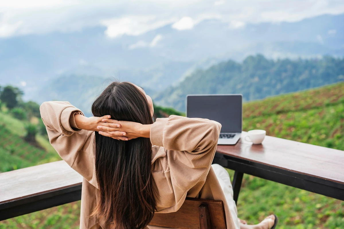 Eine junge Frau genießt ihre Workation: Sie arbeitet an ihrem Laptop, während sie die Ausblick auf die Berge genießt