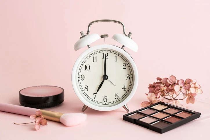 Analoger Wecker mit Make-up-Produkten vor rosa Hintergrund