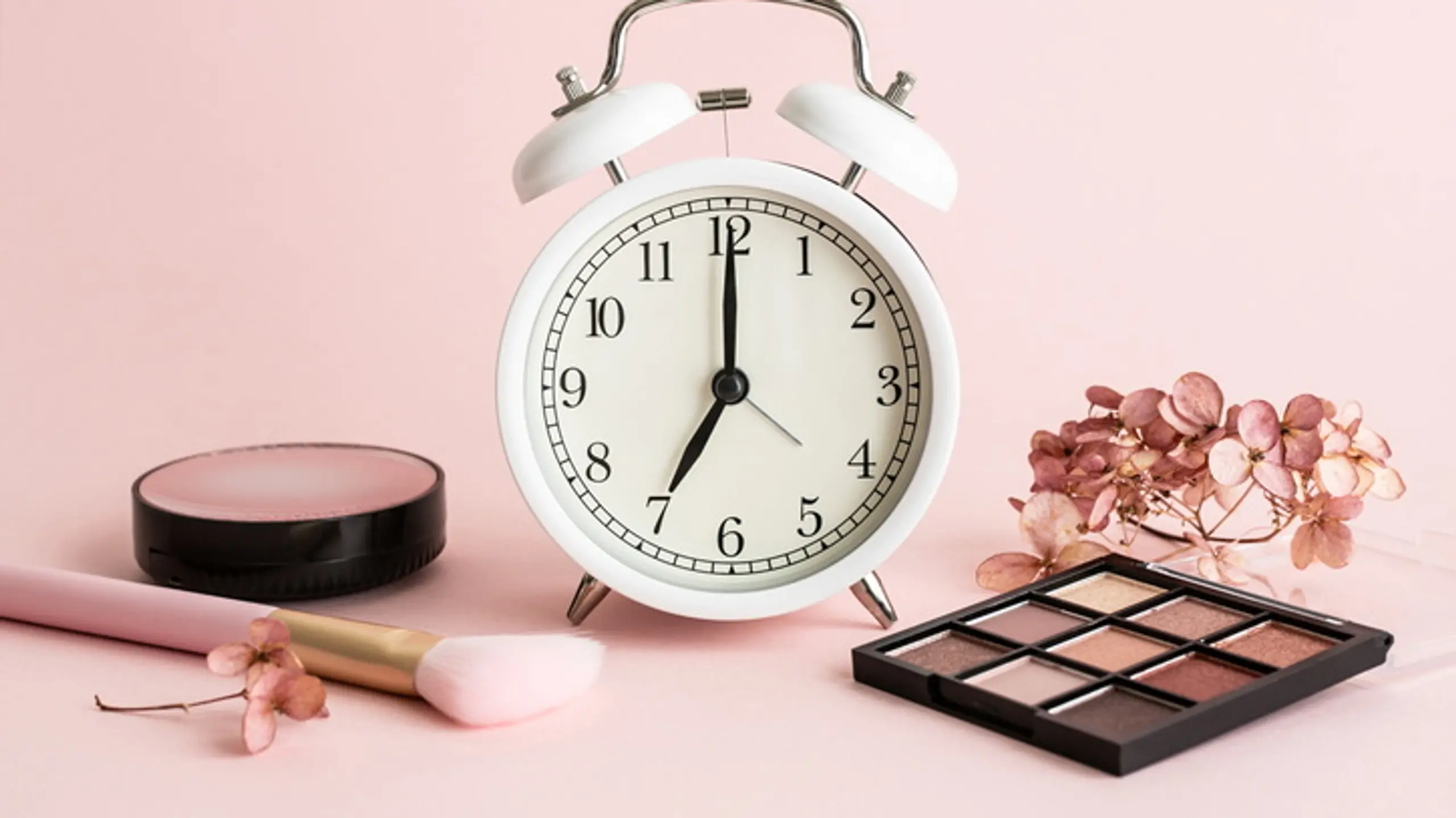 Analoger Wecker mit Make-up-Produkten vor rosa Hintergrund