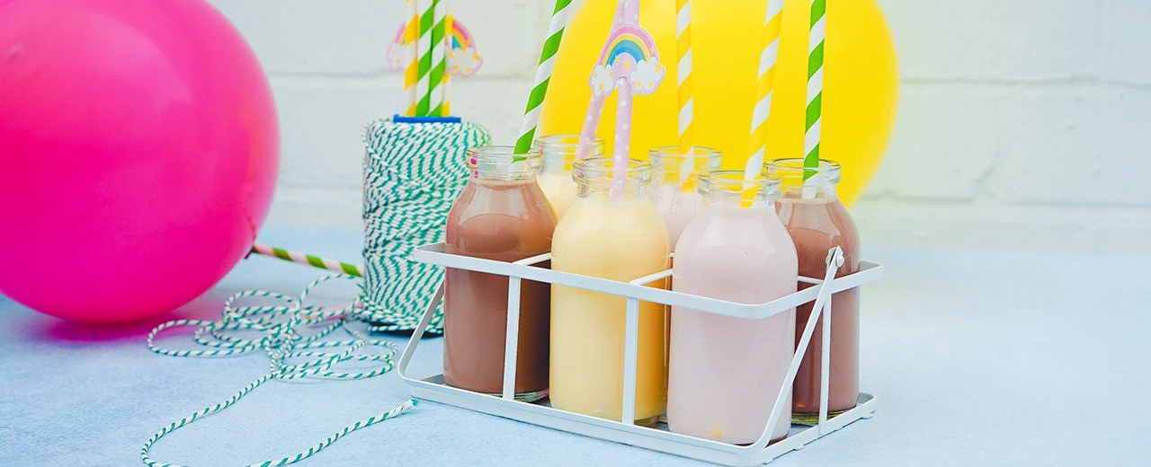 Sechs Milchflaschen mit verschiedenen Geschmacksrichtungen in einem weißen Träger, umgeben von Luftballons und bunten Strohhalmen. Perfekt für Kindergeburtstage.