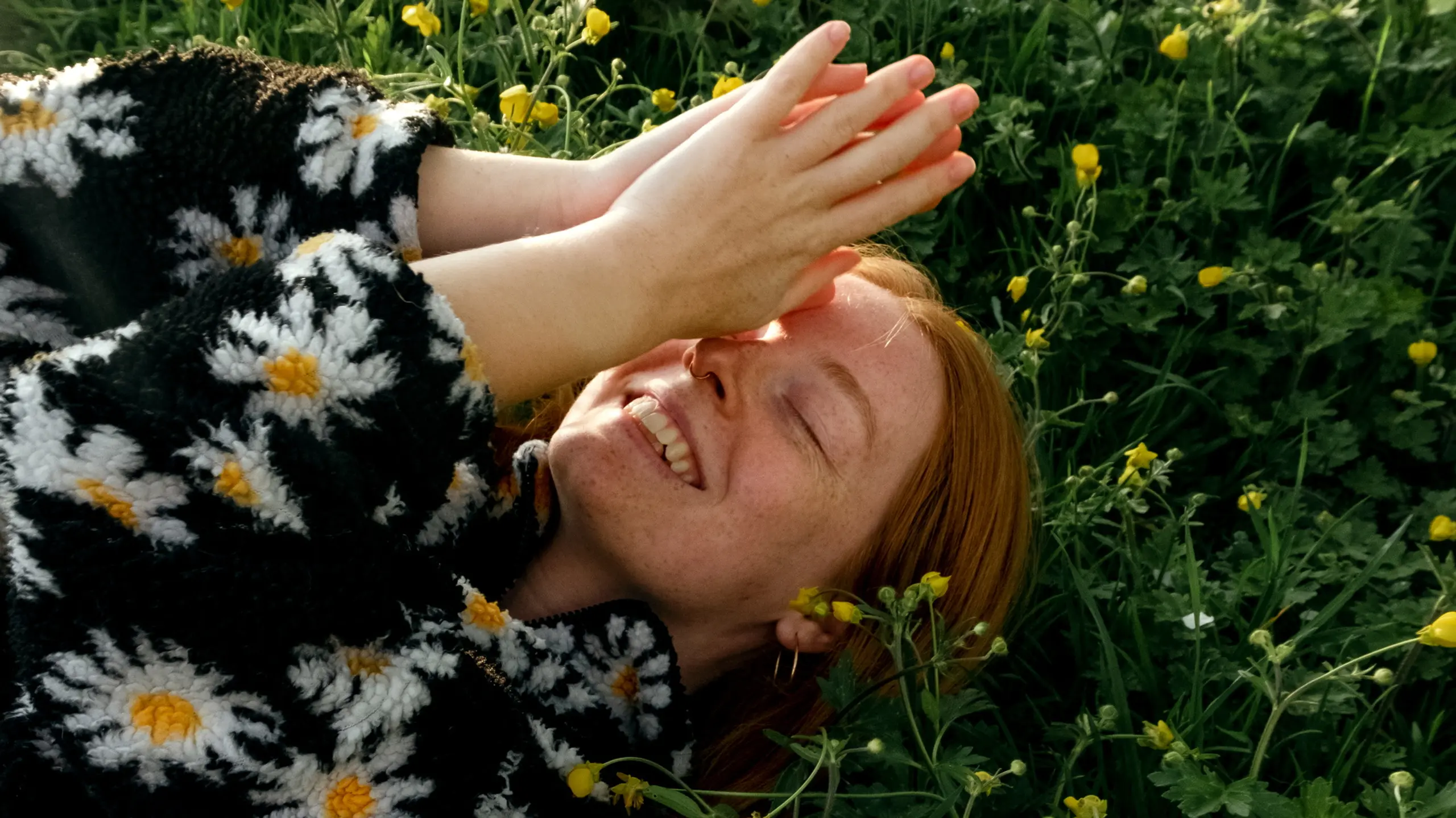 Frau mit Gänseblümchenpullover liegt in Blumenwiese und lächelt