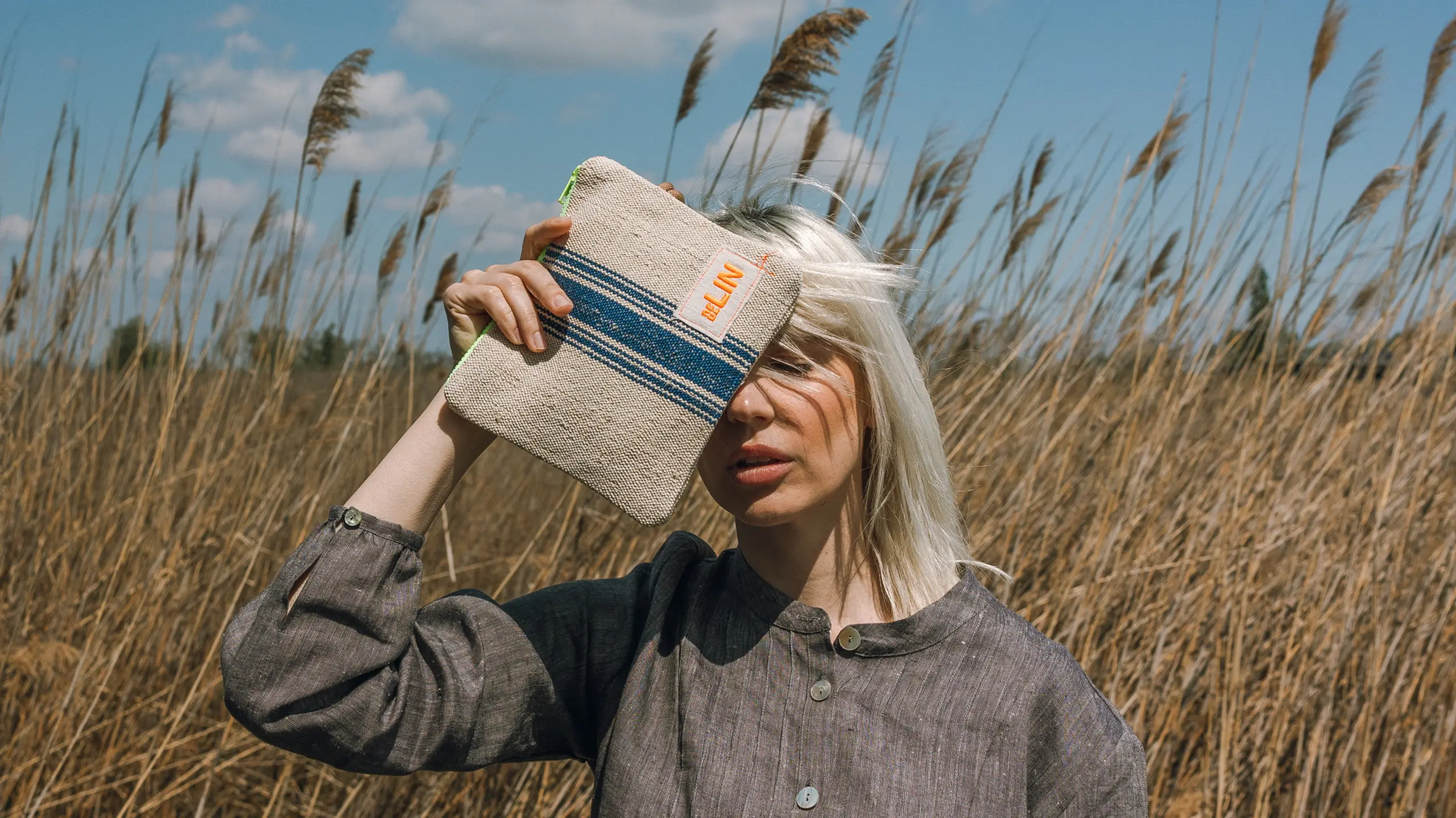 Model steht in einem Feld und hält sich eine Leinentasche vors Gesicht