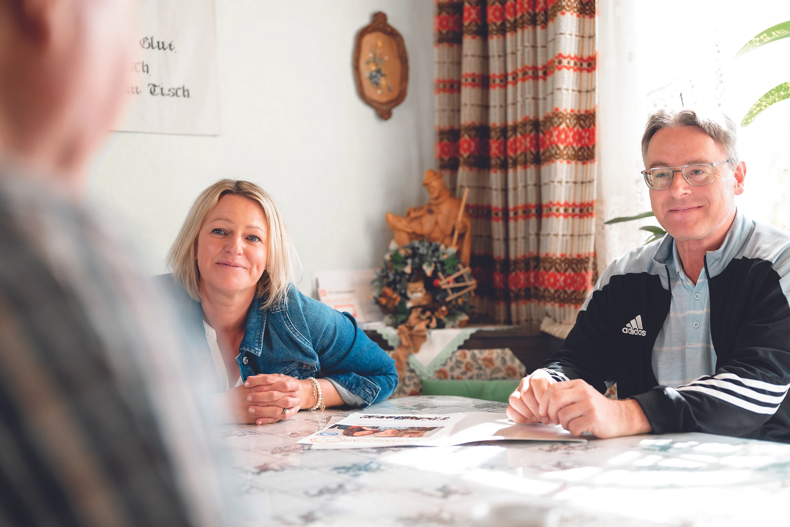 © Rene Knabl Entspanntes Paar unterhält sich an einem Küchentisch. Frau in Jeansjacke, Mann in Adidasjacke. Erstellt mit Color Factory 8.1