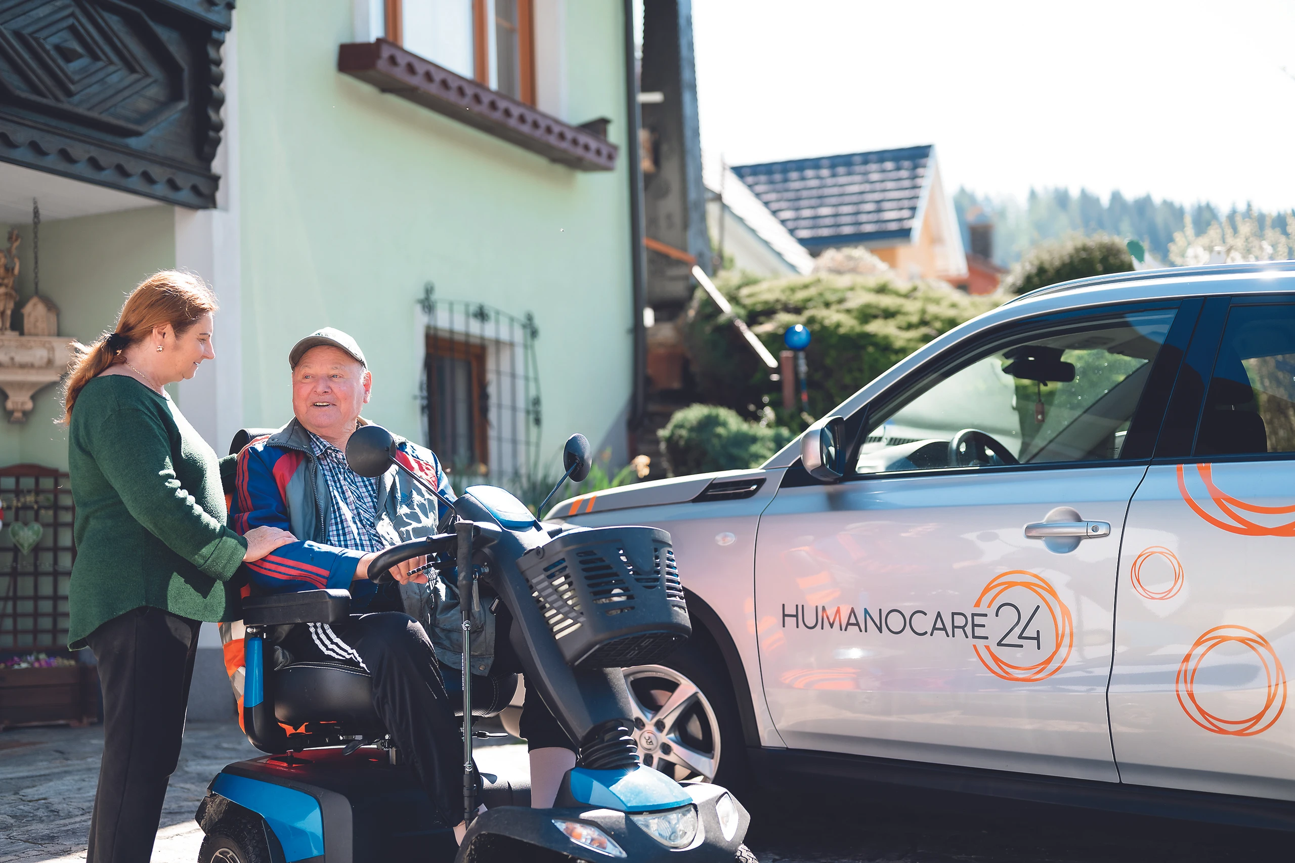Älterer Mann im Elektrorollstuhl erhält Hilfe, HumannoCare24 Fahrzeug im Hintergrund.